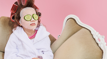 Girl with hair in curlers and cucumber slices over her eyes relaxing in a spa robe, promoting the Shimmer Hands & Feet Spritz treatment